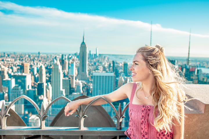 Bild zeigt eine junge Frau, die auf einer Terrasse steht und über eine Stadt blickt.