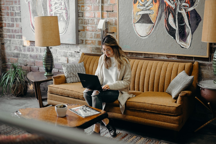 Bild zeigt eine junge Frau, die an einem Laptop auf einem Sofa arbeitet.