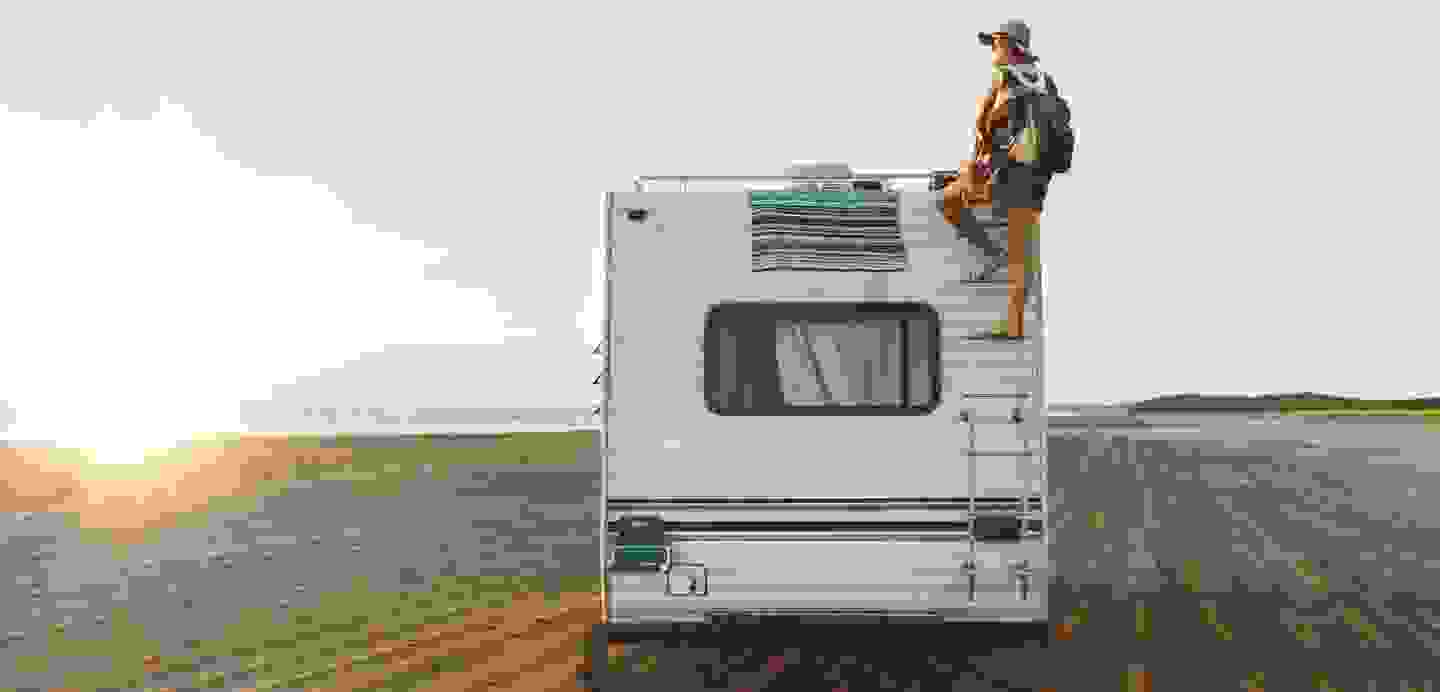 Stimmungsbild: Wohnwagen von hinten am Strand und ein Mensch, der hinten mithilfe einer Leiter auf den Wohnwagen steigt.