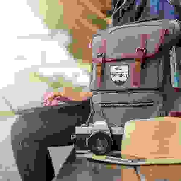 Stimmungsbild: Mensch sitzt auf einer Bank und im Vordergrund ist ein Rucksack mit TFBO-Logo, ein Fotoapparat, einen Sommerhut und ein Handy platziert.